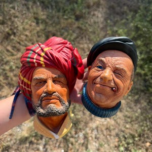 May include: Two sculpted head figurines. One features a person with a dark beard wearing a red and yellow plaid turban. The other depicts a person with a wrinkled face, wearing a black cap and a blue turtleneck, with a small cigar in their mouth.