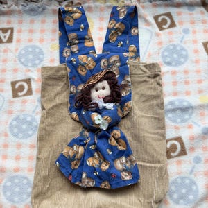 Op de afbeelding: Een beige rugzak van corduroy met een blauwe stoffen flap versierd met een patroon van teddyberen. Een kleine stoffen pop met bruin garen haar, een strooien hoed en een blauwe jurk met teddyberenprint is in het midden bevestigd.