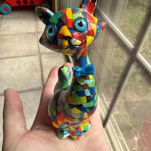 May include: A small cat figurine featuring a vibrant, multi-colored mosaic pattern across its entire body. The cat has large, round, bright blue eyes, a small black painted nose and mouth, and a solid light blue bow tie at its neck.