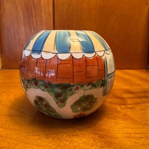 May include: A spherical ceramic vase featuring a hand-painted scene of a storefront with a blue and white striped awning, a brick-patterned wall, and green bushes with red accents. The design wraps around the rounded body of the vessel.