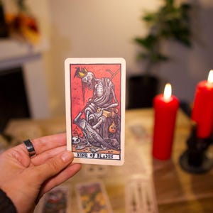 May include: A rectangular tarot card titled King of Blades featuring a skeletal figure wearing a crown and holding a scythe. The illustration uses a red background with black line work and muted tones. The card is held upright against a blurred background.