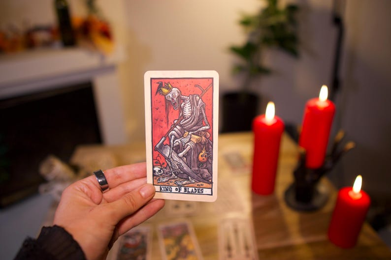May include: A rectangular tarot card titled King of Blades featuring a skeletal figure wearing a crown and holding a scythe. The illustration uses a red background with black line work and muted tones. The card is held upright against a blurred background.