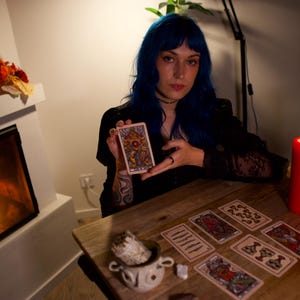 May include: A rectangular tarot card featuring a colorful, intricate illustration of a central figure with a sun-like emblem, held upright. The card displays detailed line work and a vibrant palette of gold, blue, and red tones on a white border.
