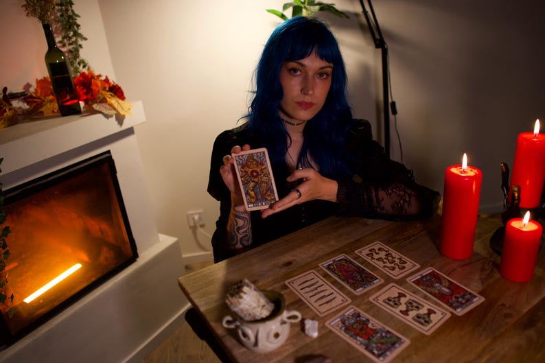May include: A rectangular tarot card featuring a colorful, intricate illustration of a central figure with a sun-like emblem, held upright. The card displays detailed line work and a vibrant palette of gold, blue, and red tones on a white border.