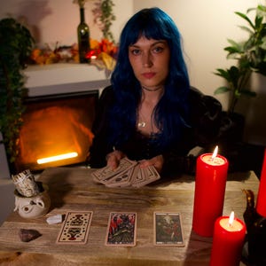 May include: A person with blue hair holds a deck of tarot cards at a wooden table. Three tarot cards are laid out face up. Two red pillar candles stand on the table, one in a hand-shaped holder. A small ceramic bowl with sage sits nearby.