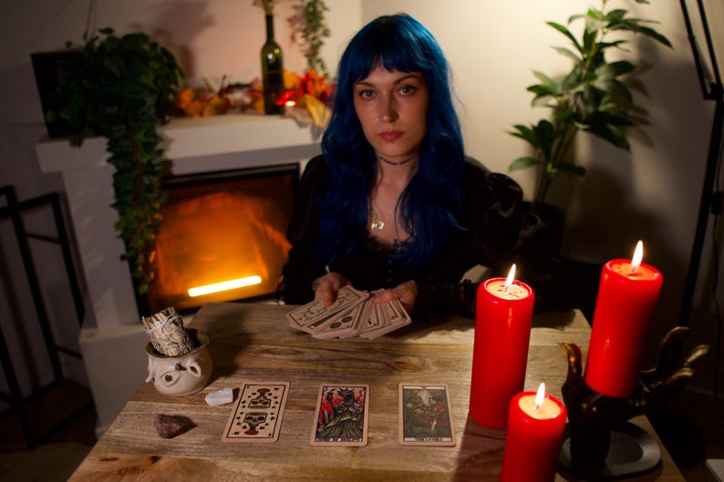 May include: A person with blue hair holds a deck of tarot cards at a wooden table. Three tarot cards are laid out face up. Two red pillar candles stand on the table, one in a hand-shaped holder. A small ceramic bowl with sage sits nearby.