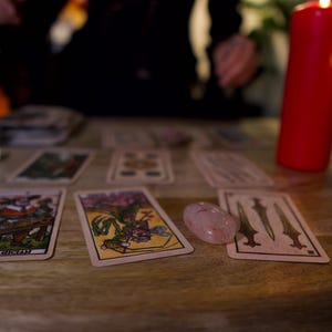 May include: A collection of tarot cards spread across a wooden surface, including The Magician card, alongside a smooth pink crystal and a tall red pillar candle. The cards feature detailed illustrations with various colors and symbolic imagery.