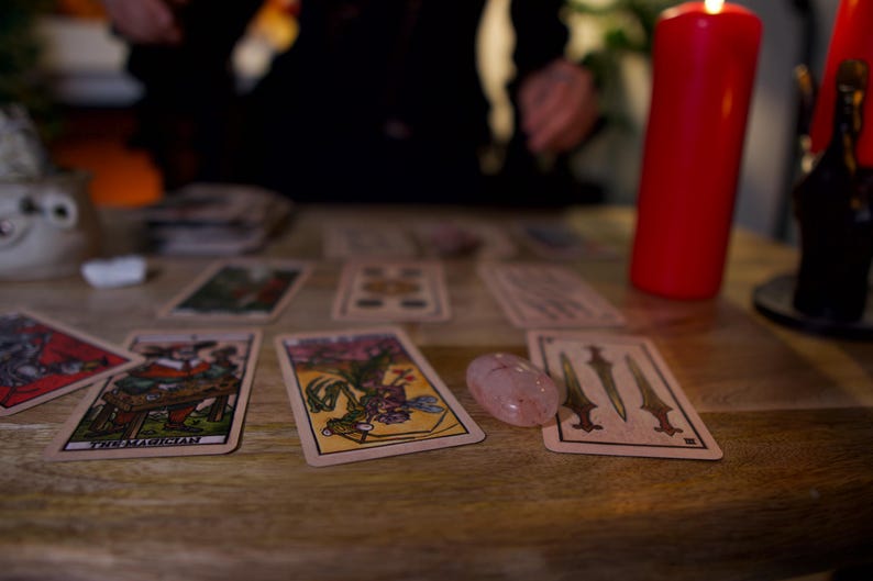 May include: A collection of tarot cards spread across a wooden surface, including The Magician card, alongside a smooth pink crystal and a tall red pillar candle. The cards feature detailed illustrations with various colors and symbolic imagery.