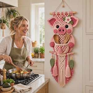 Könnte beinhalten: Ein rosa-cremefarbener Makramee-Wandbehang in Schweineform mit einer zentralen Tasche, die zwei hölzerne Küchenutensilien enthält. Das Design zeigt eine weiße Blume am Kopf, zwei grüne Blattakzente und lange dekorative Quasten am unteren Ende.