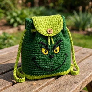 May include: A dark green crochet drawstring backpack featuring a light green flap with a single wooden button. The front displays a stitched face with yellow eyes, black eyebrows, and a wide black smile. It has matching green braided straps.