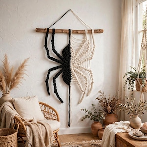 May include: A macrame wall hanging featuring a large spider design split down the middle, with the left side in black cord and the right side in cream cord. The spider is suspended from a horizontal wooden branch by a cord loop.