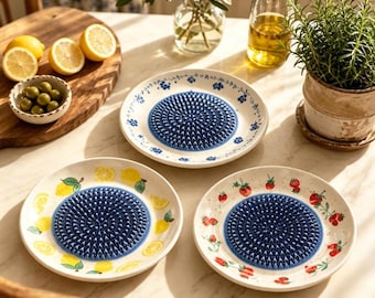 May include: Three ceramic grater plates with a central blue textured grinding surface. One plate features blue floral patterns, one has yellow lemon illustrations, and one displays red strawberry motifs, all arranged on a white tabletop surface.