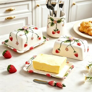 May include: Two white ceramic butter dishes with lids, each featuring a hand-painted strawberry and flower vine pattern and a sculpted strawberry knob. Includes a matching utensil holder with cutlery, a butter knife with a strawberry handle, and fresh strawberries.