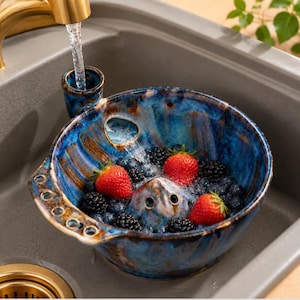 May include: A round, blue, glazed ceramic berry colander with a built-in water funnel and drainage holes. The bowl is filled with fresh strawberries and blackberries being rinsed under a stream of water from a gold-toned kitchen tap.
