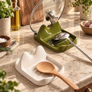May include: Two ceramic spoon rests with a raised back support for holding pot lids. One rest is olive green and holds a metal ladle and a glass lid, while the other is white and holds a wooden spoon. Both feature a grooved base for drainage.
