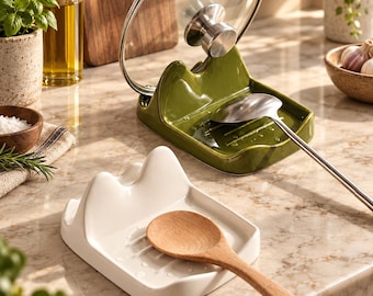 May include: Two ceramic spoon rests with a raised back support for holding pot lids. One rest is olive green and holds a metal ladle and a glass lid, while the other is white and holds a wooden spoon. Both feature a grooved base for drainage.
