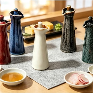 Puede incluir: Cinco dispensadores de salsa de soja de cerámica en rojo, azul, blanco, gris moteado y verde moteado. Cada botella presenta un cuello cónico, un pequeño pico vertedor y una tapa a juego, dispuestos sobre una alfombrilla gris con cuencos.
