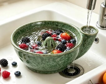 May include: A sage green ceramic berry colander with a speckled finish and a central cone, filled with water and fresh berries including strawberries, blackberries, raspberries, and blueberries, positioned under a running kitchen tap.