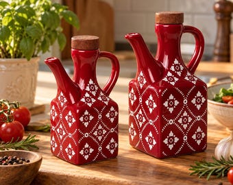 May include: Two square-shaped red ceramic oil and vinegar cruets with white geometric floral patterns and brown cork stoppers. Each cruet features a curved handle and a narrow pouring spout, showcasing a glossy finish on the ceramic surface.