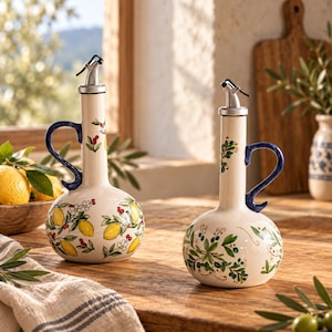 Può includere: Due oliere in ceramica color crema con manici blu e beccucci argentati. Un contenitore presenta un motivo a limoni gialli e fiori bianchi, mentre l'altro mostra un disegno di foglie verdi e bacche blu sui loro corpi arrotondati.