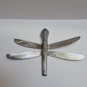 May include: A dragonfly-shaped sculpture constructed from silver-toned metal cutlery. The body consists of a single ornate handle with floral engravings, while the four wings are formed by four individual serrated dinner knives.