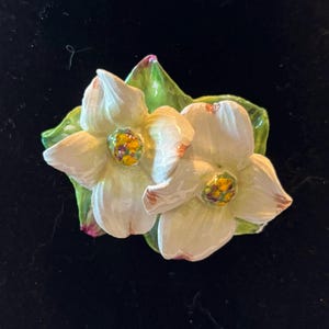 May include: A ceramic brooch featuring two white dogwood flowers with four petals each, displaying subtle pink-tinged edges and textured yellow and purple centers, set against a backdrop of glossy green leaves with visible veining and depth.