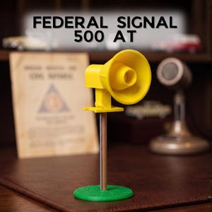 Könnte beinhalten: Ein gelbes Federal Signal 500 AT Sirenenmodell, montiert auf einem dünnen braunen Stab mit einem grünen runden Sockel. Die Sirene hat eine markante konische Hornform. Der Text FEDERAL SIGNAL 500 AT steht in schwarzen Buchstaben darüber.