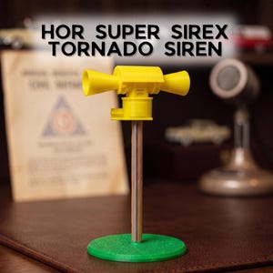 May include: A yellow HOR SUPER SIREX TORNADO SIREN model mounted on a thin wooden pole with a silver vertical accent, supported by a round green base. The siren features two horizontal flared horns extending from a central cylindrical housing.