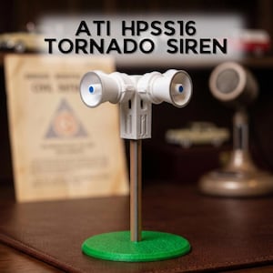 May include: A white ATI HPSS16 tornado siren model featuring two megaphone-style speakers mounted on a central pillar. The unit is supported by a thin vertical pole attached to a circular green base. The text ATI HPSS16 TORNADO SIREN is visible.