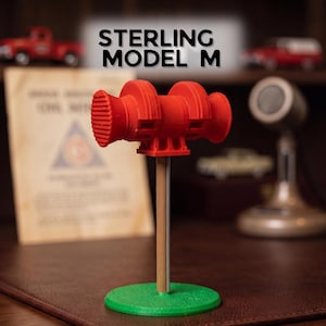 May include: A red Sterling Model M siren replica mounted on a vertical wooden dowel attached to a circular green base. The siren features a cylindrical body with ribbed ends and a central mounting bracket, displaying the text STERLING MODEL M above.