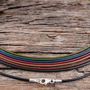 May include: A set of leather cord necklaces in various colors, including black, brown, red, blue, and green. The cords are arranged in a circle on a wooden surface. A silver clasp is attached to the black cord.