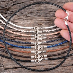 A hand holding a black braided leather necklace with a silver clasp, surrounded by various colored braided leather necklaces on a wooden surface.