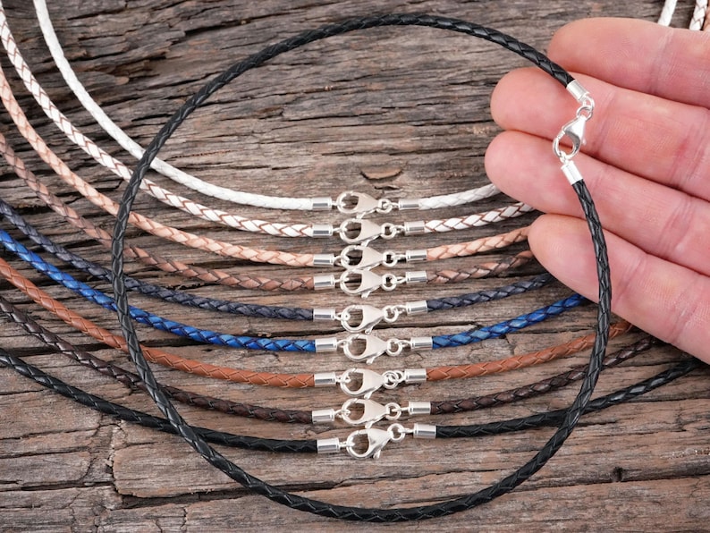 A hand holding a black braided leather necklace with a silver clasp, surrounded by various colored braided leather necklaces on a wooden surface.