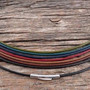 May include: A variety of leather cords in different colors, including black, brown, red, blue, and green, are arranged in a rainbow pattern. The cords are all the same width and have a silver clasp.