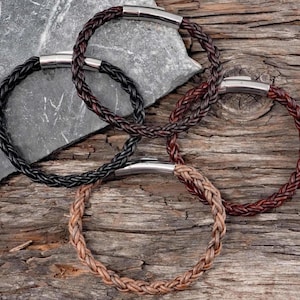 May include: Four braided leather bracelets in black, brown, and tan, each with a silver-tone clasp. The bracelets are arranged on a weathered wooden surface, with a gray stone in the background. The bracelets are a stylish accessory.