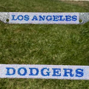 May include: A rectangular license plate frame covered in clear, shimmering rhinestones. The top bar features the text LOS ANGELES in blue rhinestones, while the bottom bar displays the text DODGERS with the LA logo in blue rhinestones.