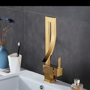 May include: A gold-colored bathroom faucet with a tall, vertical, curved spout and a square base. The faucet features a single side-mounted lever handle for water control. The design is metallic with a reflective finish, positioned on a white sink.