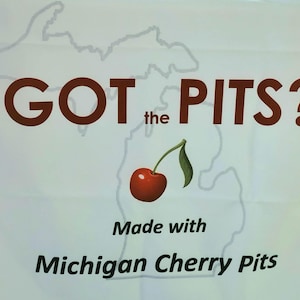 May include: A white rectangular sign featuring a light gray outline of the state of Michigan. The text reads GOT the PITS? in brown, with a red cherry illustration in the center. Below, the text reads Made with Michigan Cherry Pits in black.
