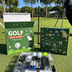 Op de afbeelding: Een golf-adventskalenderset met een groene doos met de tekst GOLF ADVENT CALENDAR MERRY CHRISTMAS, een tweede groene doos met genummerde deurtjes van 1 tot 24, en een doorzichtige plastic tray met diverse golfaccessoires en witte tees.