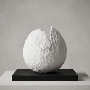 Op de afbeelding: Een witte, bolvormige sculptuur met een ruw, getextureerd oppervlak en een grillige verticale scheur die vanaf de bovenkant naar beneden loopt. Het object is bevestigd op een platte, rechthoekige zwarte voet, wat een scherp contrast vormt.