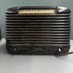 Könnte beinhalten: Ein schwarzes Vintage-Radio der Marke Sparton mit horizontal geripptem Gehäuse und einer Skala an der Oberseite mit Zahlen von 55 bis 16. Auf der Vorderseite befindet sich der Schriftzug Sparton sowie zwei runde Drehknöpfe an den unteren Ecken.