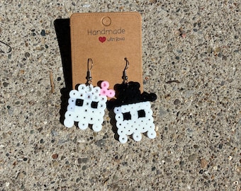 May include: A pair of ghost-shaped earrings crafted from white and black plastic beads. One ghost features a small pink bow, while the other wears a black top hat. The earrings are attached to a brown cardboard display card labelled Handmade with love.