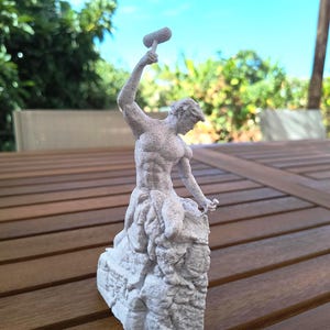 Op de afbeelding: Een wit, getextureerd beeldje dat een gespierd figuur in een dynamische houding weergeeft, met een hamer hoog boven het hoofd geheven terwijl een kleine beitel tegen een blok steen wordt gehouden. Het beeld heeft een ruwe, korrelige afwerking.