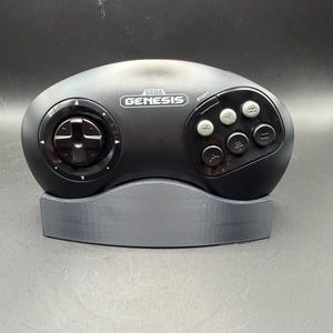 Op de afbeelding: Een zwarte Sega Genesis videogamecontroller met een cirkelvormige richtingspad aan de linkerkant en zes grijze actieknoppen met de labels A, B, C, X, Y en Z aan de rechterkant. De controller rust op een gebogen, donkergrijze plastic displaystandaard.