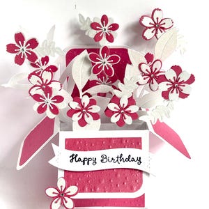 May include: A handmade birthday card with a pop-up floral design. The card is a bright pink with white details, including paper flowers and leaves. The words "Happy Birthday" are written on a white banner.