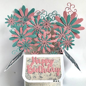 May include: A pop-up card with a 3D design of pink and teal flowers. The card is open and says "Happy Birthday" inside.
