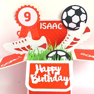 May include: A red and white pop-up card with a football theme. The card is open and shows a football, a pair of football boots, and a green grass field. The card says "Happy Birthday" and "Isaac" on the front. The card also has a red ribbon with a white circle that says "9" on it.