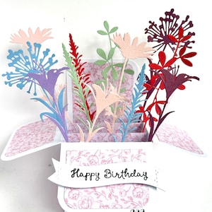 May include: A pop-up birthday card with paper flowers in pink, purple, red, and blue. The card has a white banner with the words "Happy Birthday" and a floral pattern inside. The card is a unique gift.