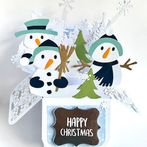 May include: A 3D pop-up greeting card featuring three paper snowmen with orange carrot noses, dark blue hats, and scarves. The card includes green paper pine trees, white snowflake cutouts, and a central plaque with the text Happy Christmas.