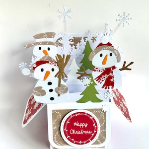 May include: A three-dimensional pop-up greeting card featuring three snowmen, green pine trees, and white snowflakes. The base is a square box with a red circular label that reads Happy Christmas in white script, surrounded by a white border.